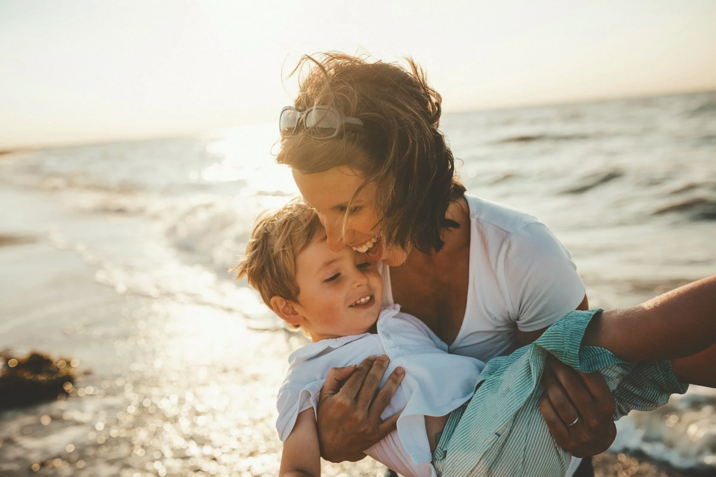 A laughing mother dips a joyous child playfully in front of the ocean, as the sun shines from behind her.